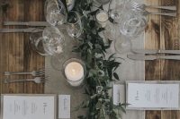 a chic winter wedding tablescape with a grey table runner and linens, candles, a greenery runner and elegant cutlery