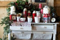 a cozy Christmas hot chocolate bar with evergreens, plaid elements, a chalkboard sign, a Christmas arrangement