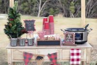 a cozy rustic hot chocolate bar with plaid banners, metal buckets, a mini Christmas tree and some cups