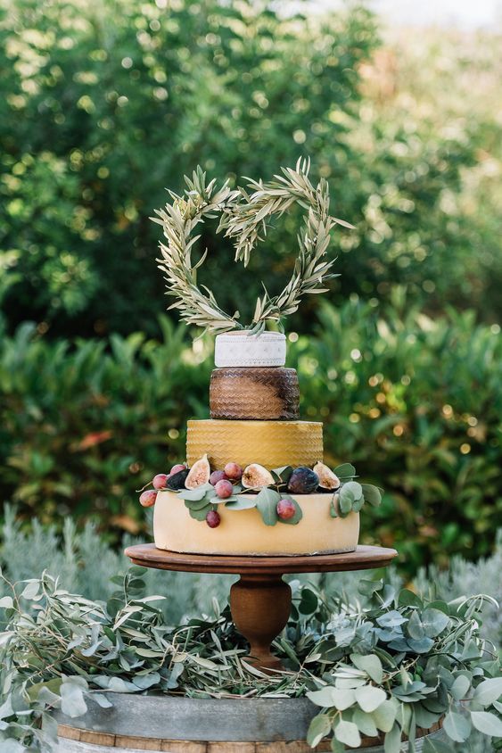 a fabulous cheese wheel wedding cake topped with greenery, figs, grapes and with a greenery heart-shaped topper for a summer wedding