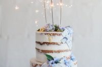 a naked wedding cake decorated with blue flowers and blue marble tiles, with sparklers on top that give a party feel to the cake