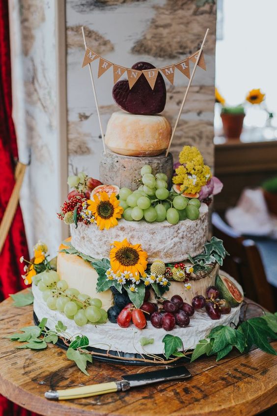 a rustic cheese wheel wedding cake topped with super bold blooms, grapes, figs and tomatoes plus a burlap banner on top