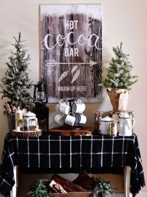 a stylish hot chocolate bar with snowy trees, some metal mugs, lanterns, a snowy sign on the wall and a crate with firewood