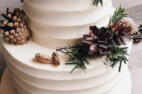 a textural white wedding cake topped with evergreens, nuts and pinecones and cinnamon sticks