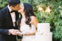 a white buttercream wedding cake decorated with neutral blooms, greenery and sparklers on top us a stylish and cool solution for a wedding