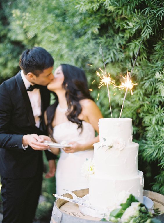 a white buttercream wedding cake decorated with neutral blooms, greenery and sparklers on top us a stylish and cool solution for a wedding