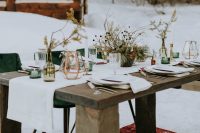 a winter boho wedding table with a white runner, green candleholders and vases, a copper candle lantern and burgundy linens, dried blooms