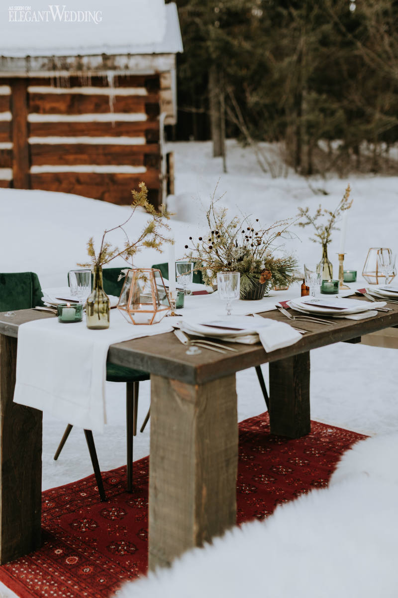A winter boho wedding table with a white runner, green candleholders and vases, a copper candle lantern and burgundy linens, dried blooms
