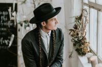 a winter groom outfit with grey pants, a shirt, a blazer, a black hat and long braids for a boho touch
