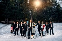 a groom and his groomsmen in black tuxedos and with their colorful snowboards and skis