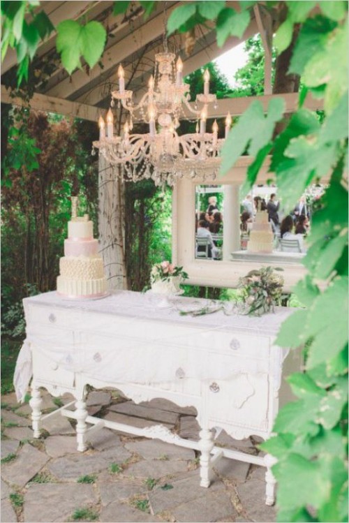a white vintage dresser with a mirror, a crystal chandelier and blooms and a couple of delicious wedding cakes served is a refined idea