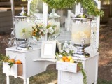 a white vintage dresser with a large mirror, greenery runners and citrus, used as a wedding drink station, with lemonade tanks