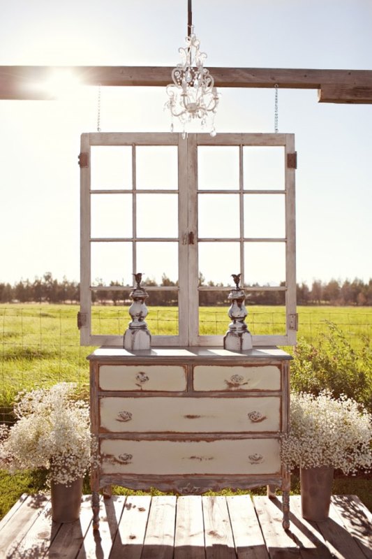 a chic vintage wedding space with a window frame, a crystal chandelier, a vintage neutral dresser, baby's breath arrangements in buckets and candleholders