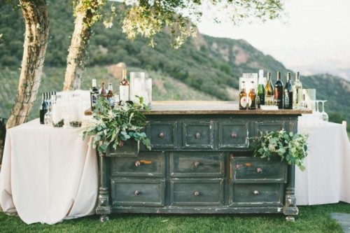 a large shabby chic vintage dresser used as a wedding bar, decorated with greenery and blooms and with additional tables on both sides