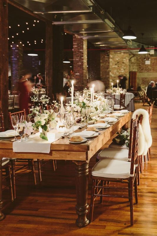 an industrial wedding venue with brick walls, pillars, hanging lights, candles, potted greenery and white blooms and comfy chairs