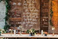 an industrial wedding venue with concrete walls, a pallet decoration, a greenery garland, a long table with blooms and candles is very chic