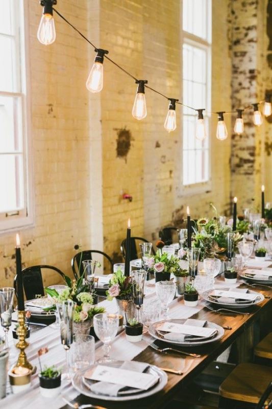 a neutral industrial wedding venue with light-colored brick walls, hanging lights, greenery, pink blooms, black candles and potted greenery as favors