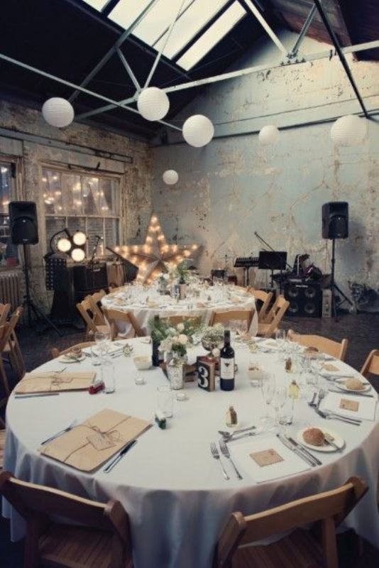 an industrial wedding venue with shabby chic walls, exposed metal beams is softened with marquee lights and paper lamps plus neutral blooms