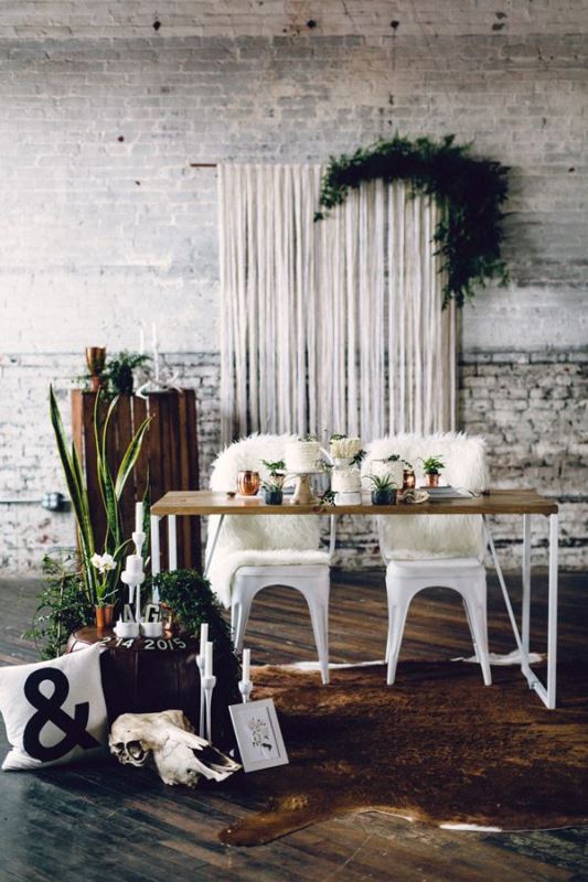 an industrial wedding venue with white brick walls, a wooden pallet, a table with metal chairs and faux fur, a macrame hanging with greenery and some potted plants