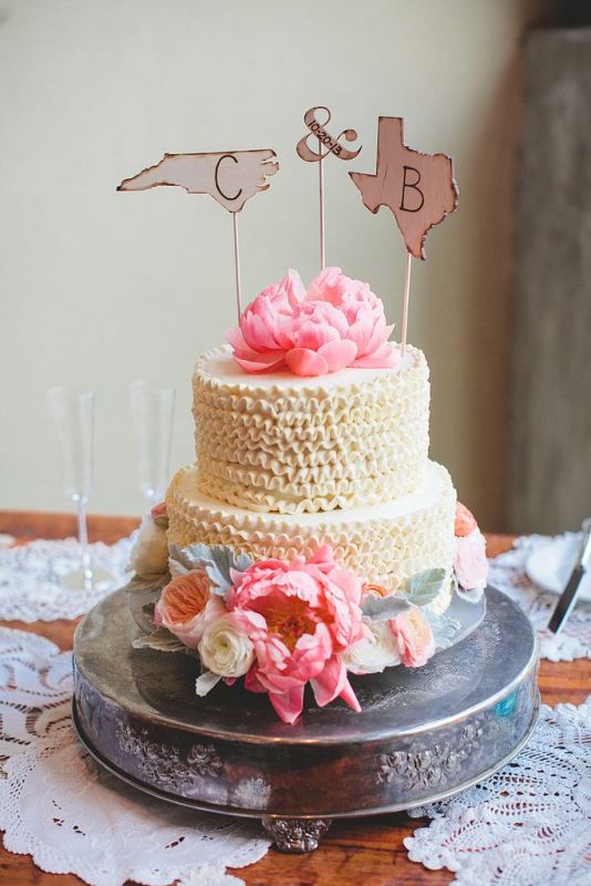 a ruffled wedding cake decorated with fresh pink blooms and topped with map parts