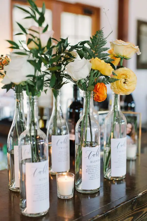 a beautiful cluster wedding centerpiece of wine bottles, bright blooms and greenery and some candles is a lovely idea for many weddings