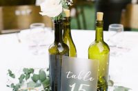 a beautiful modern wine bottle wedding centerpiece of green bottles with white blooms and greenery, LED lights, greenery and a frosted glass table number