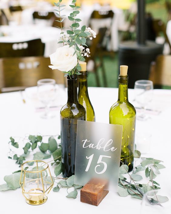 a beautiful modern wine bottle wedding centerpiece of green bottles with white blooms and greenery, LED lights, greenery and a frosted glass table number