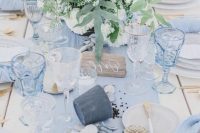 a blue beach wedding table with a light blue runner, glasses, napkins, neutral blooms, pebbles and seashells