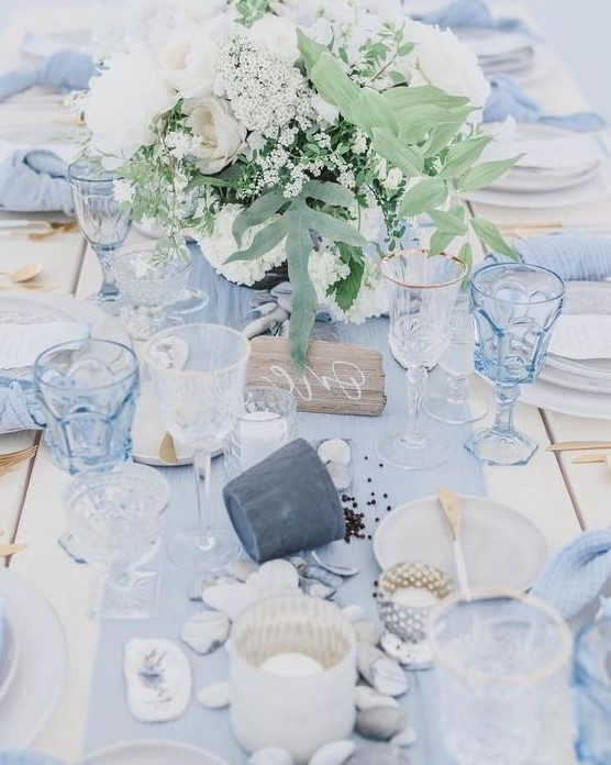 a blue beach wedding table with a light blue runner, glasses, napkins, neutral blooms, pebbles and seashells