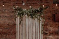 a boho industrial wedding ceremony space with a fringe backdrop, greenery on top, floating candles and a boho rug