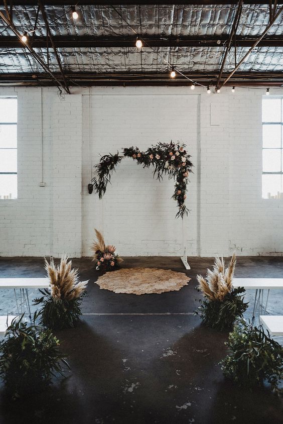 a boho industrial wedding ceremony space with a white arch decorated with greenery and pink blooms, greenery and pampas grass