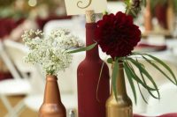 a bright and relaxed wedding centerpece of a wood slice, beer and a wine bottle, baby’s breath and a burgundy bloom, greeneyr and a table number