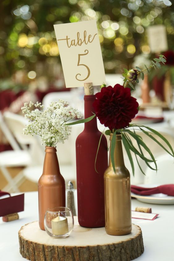 a bright and relaxed wedding centerpece of a wood slice, beer and a wine bottle, baby's breath and a burgundy bloom, greeneyr and a table number
