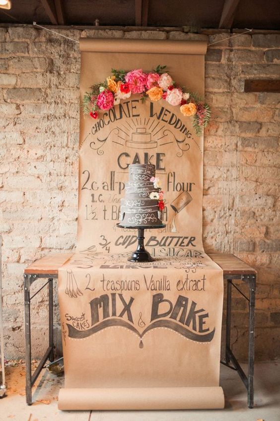 a cake table with a kraft paper backdrop with bold blooms and a chalkboard wedding cake decorated with blooms are a cool and creative idea