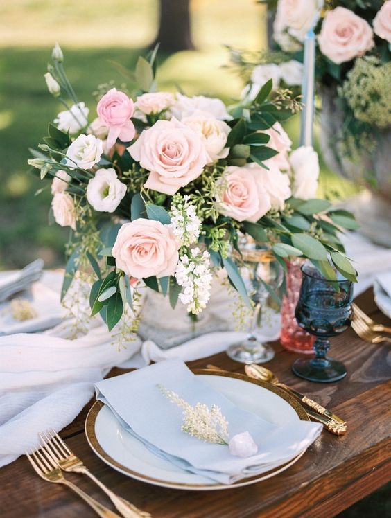 a chic pastel wedding tablescape with a serenity blue table runner and napkins, blush roses and greenery, navy glasses and gold cutlery