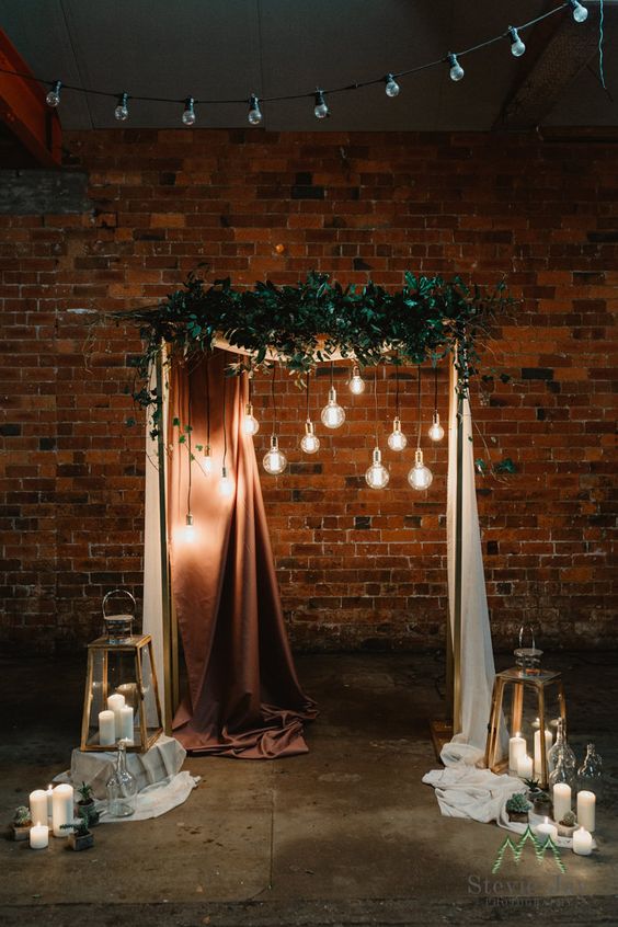 a chic wedding arch decorated with greenery, bulbs, elegant curtains, candles and potted cacti