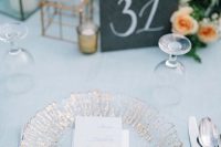 a chic wedding table setting with a serenity blue tablecloth, a gilded plate, peachy blooms and neutral napkins