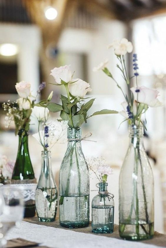 a delicate cluster wedding centerpiece of clear bottles, white blooms, greenery placed on a stand is a lovely idea to try