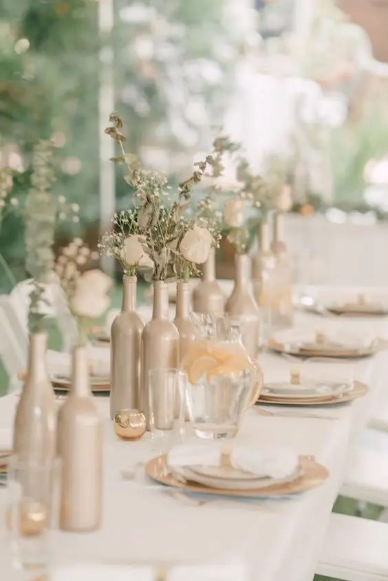 a delicate wedding centerpiece of wine bottles painted tan, white blooms and greenery is a very cool idea to try