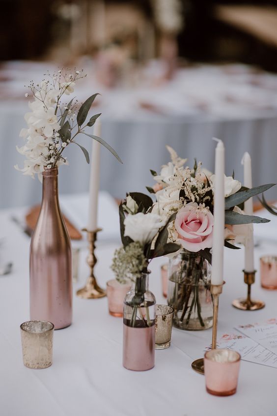 a lovely wedding centerpiece of wine bottles completely and partly painted copper, white and pink blooms and greenery and tall and thin candles