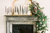 a mirror in a refined frame, tall black candles in gold candleholders and a lush greenery and bold bloom garland cascading down from the mantel