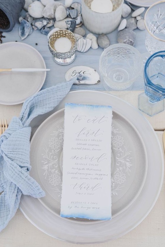 a powder blue table runner, napkin, an ombre menu, pebbles and seashells for a chic coastal look