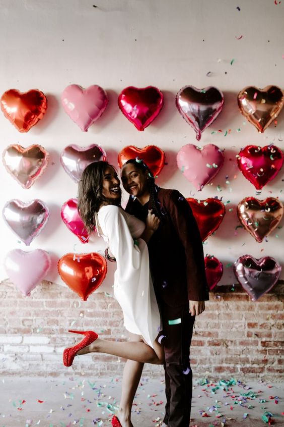 a pretty Valentine engagement pic with a heart balloon wall, bride-to-be wearing red polka dot shoes