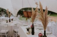 a pretty wedding centerpiece of dark wine bottles with pampas grass, some white blooms and candles plus king proteas is a very unsuaul boho solution