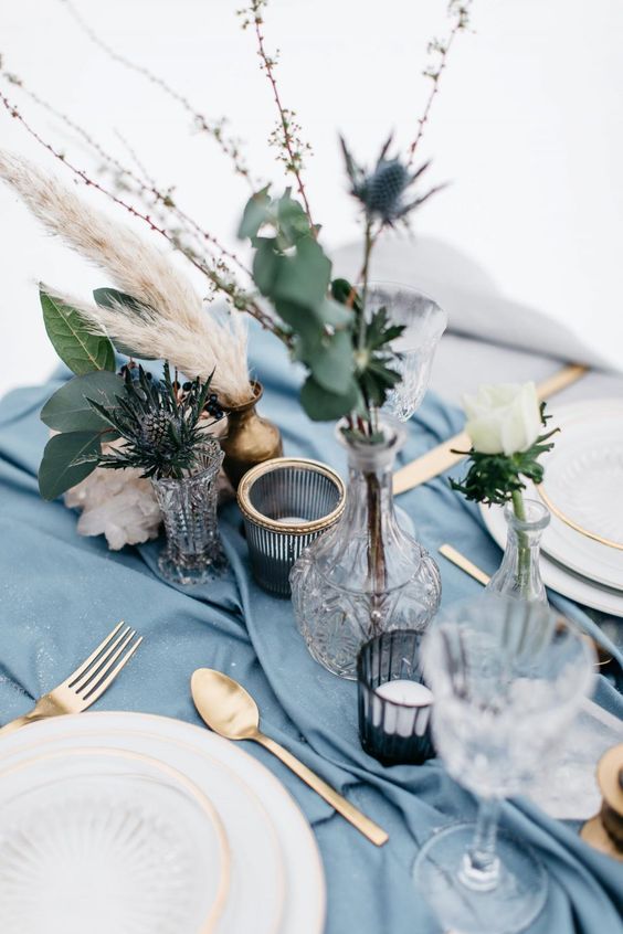 a refined wedding tablescape with a serenity blue table runner, thistles and white blooms, grasses, gold cutlery and gold rim plates