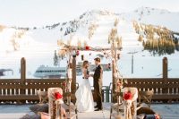 a rustic ski resort wedding ceremony space with a wooden arch, red blooms and pinecones and benches with pillows