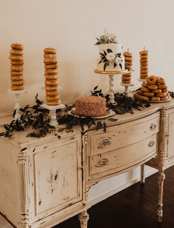 a shabby chic dresser with a greenery runner, a couple of lovely wedding cakes and glazed donuts is a chic and cool solution