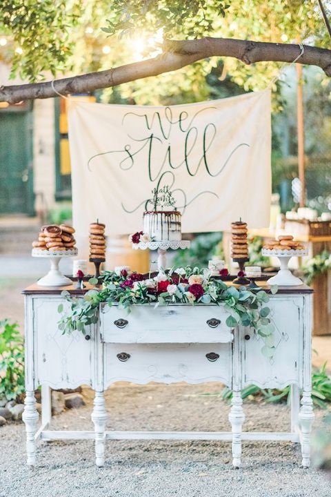 a shabby chic dresser with greenery, neutral and burgundy blooms, a wedding cake and donues plus a banner in the backdrop