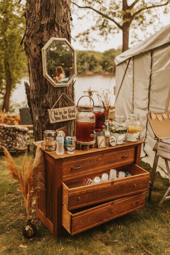 a stained vintage dresser styled as a wedding drink bar, with a mirror, dried grasses and various drinks and glasses
