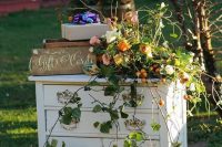 a vintage dresser with lovely wedding decor – a floral and greenery arrangement with fruit, wooden boxes with petals and a sign